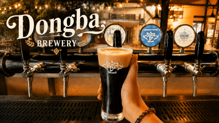 Ly bia đen có lớp bọt màu nâu, cầm phía trước dãy vòi bia và logo Đông Ba Brewery