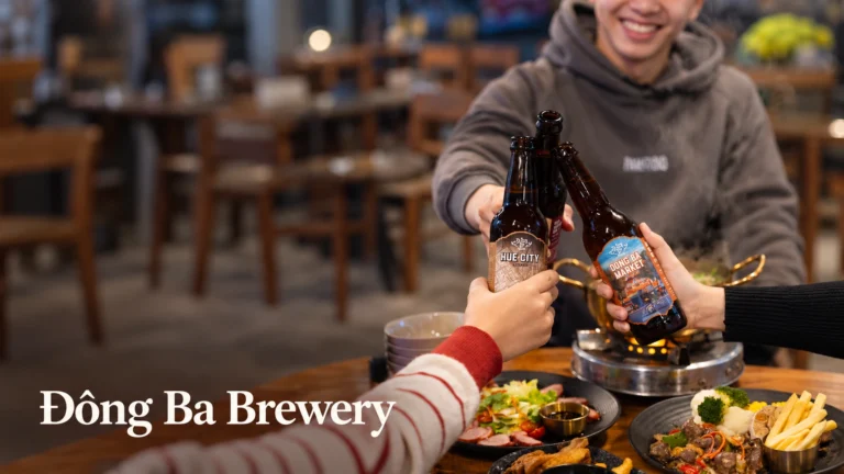 Ba người chạm chai bia trên bàn ăn có món ăn và nồi lẩu, kèm dòng chữ Đông Ba Brewery.