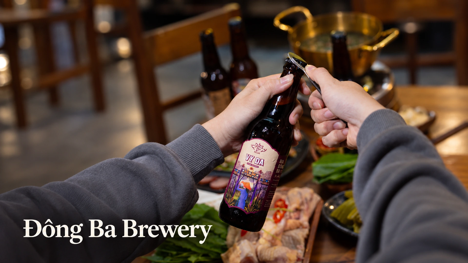Đôi tay dùng dụng cụ mở nắp chai bia Vỹ Dạ trên bàn ăn, kèm dòng chữ Đông Ba Brewery.
