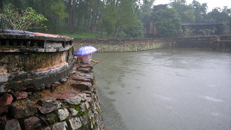 Huế when it rains: a pond beside old stone walls and a person beneath an umbrella